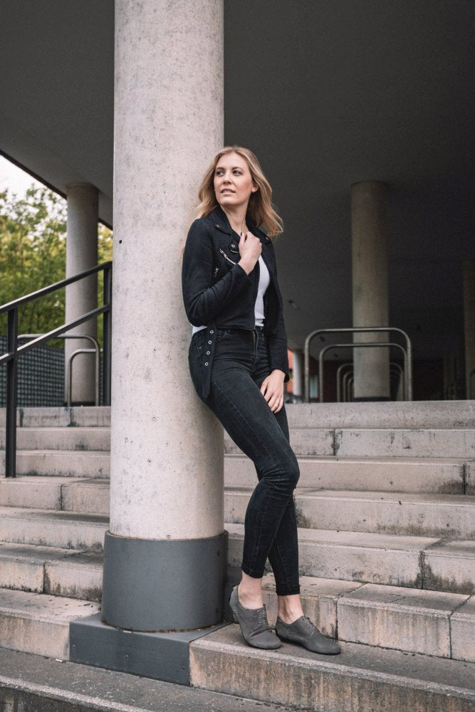 En kvinde i en grå IQON Velours-jakke og jeans fra ZAQQ læner sig mod en søjle på trinene og legemliggør perfekt business-model-stilen. Med en hånd i lommen og den anden i hendes hår udstråler hun selvtillid. Baggrunden med søjler og diskret grønt fuldender scenen.