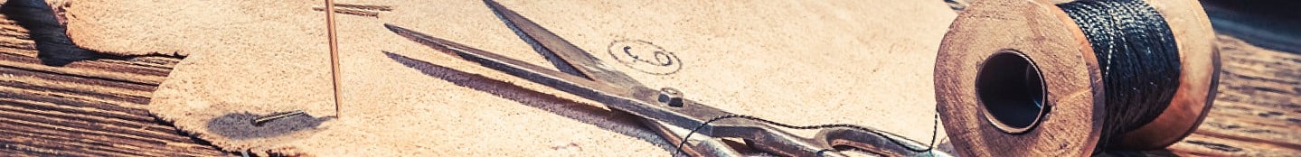 Nahaufnahme einer Vintage-Schere, die auf einem Stück rustikalem Leder neben einer Spule mit dickem schwarzen Faden liegt. Eine große Nadel durchsticht das Leder, und die Szene ist auf einer verwitterten Holzoberfläche angesiedelt, was eine antike, handgefertigte Atmosphäre ausstrahlt.