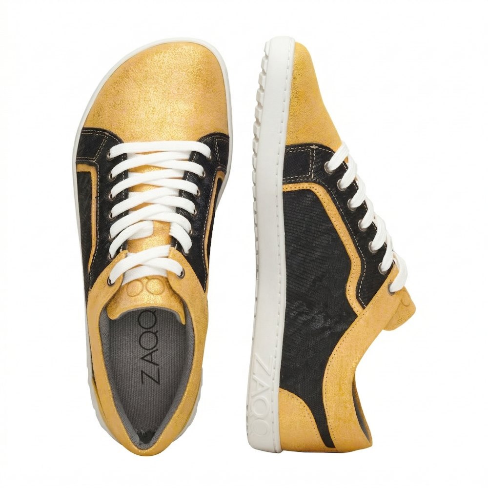 Ein Paar gelb-schwarze Sneaker mit weißen Schnürsenkeln. Ein Schuh steht aufrecht und zeigt die Seitenansicht, während der andere die Sohle und die Draufsicht zeigt. Der Markenname ZAQQ sticht auf der Seitensohle hervor und bietet unvergleichlichen Komfort, der an das Gefühl eines Barfußschuhs erinnert, bekannt als QODY Gold.