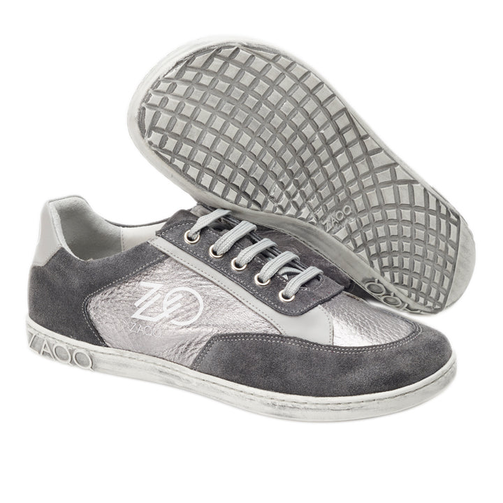 Grau-weiße Sneaker aus Veloursleder mit strukturiertem Muster an den Seiten. Ein Schuh steht aufrecht und zeigt das ZAQQ-Logo an der Seite und der Zunge, während der andere auf der Seite liegt und seine Sohle mit Waffelmuster präsentiert. Produktname: OQTAV Grey von ZAQQ.